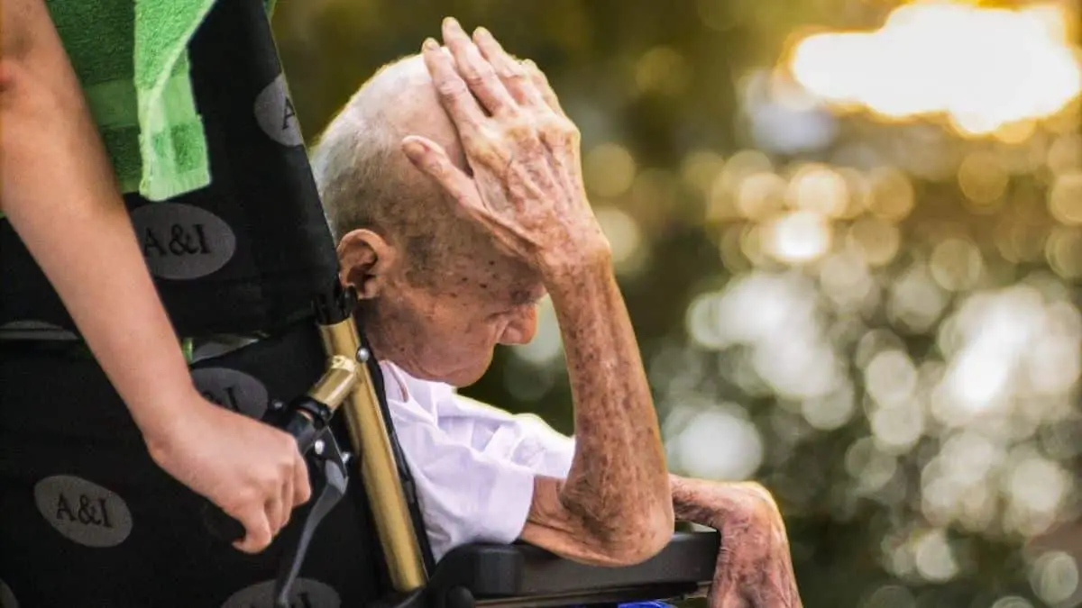 Side view of thin elderly man sitting in wheelchair with his hand on his head.