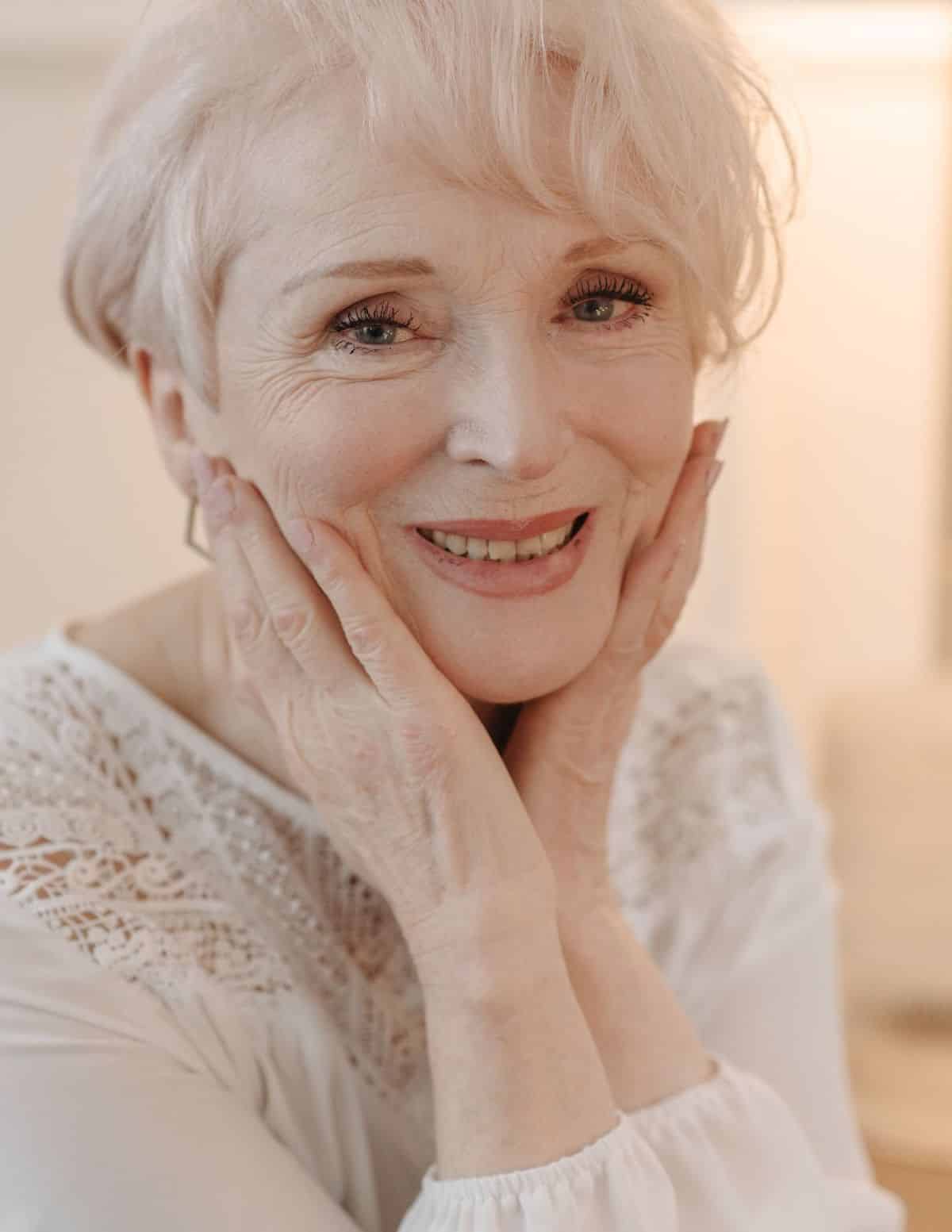 Photo of older white female with short white hair and white shirt holding her hands on her face smiling.