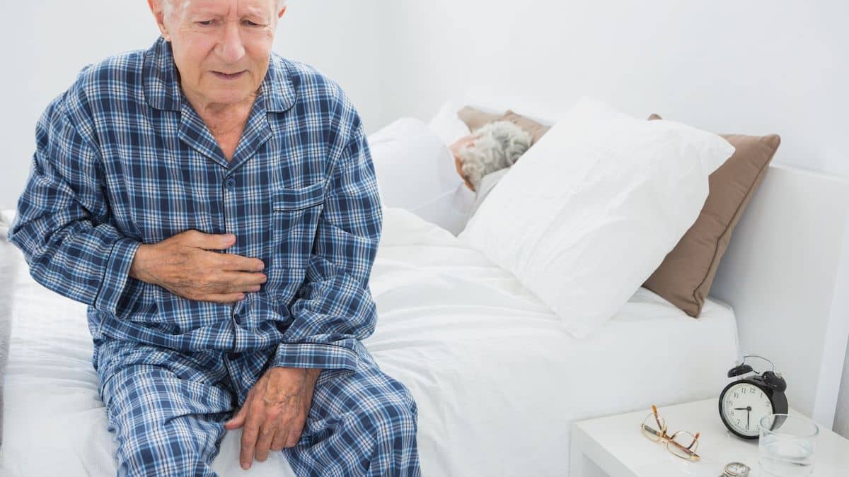 Elderly man in blue plaid pajamas sitting on the side of the bed holding his stomach in pain while a female is sleeping in the bed facing the opposite way.