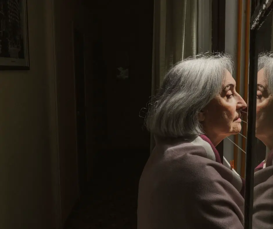 Photo of older woman standing in hallway looking out a window looking melancholy.