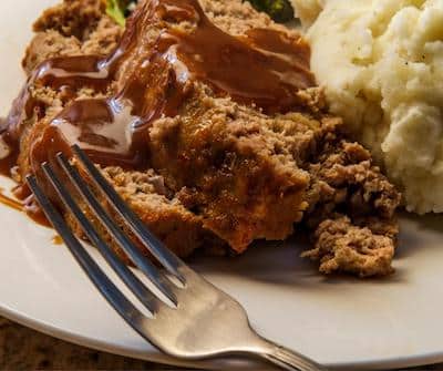Meatloaf with gravy and mashed potatoes on white plate with fork sitting in front of meal.