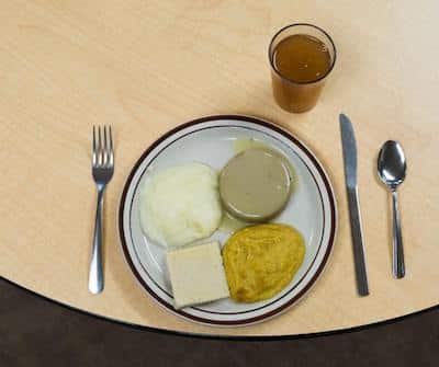 Pureed meal on white and brown plate with fork on the left and spoon and knife on the right and beverage above on a table. 