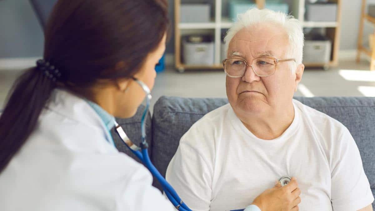 Elderly man with white hair and glasses getting his heart and lungs listened to with a stethoscope by a female health professional in a lab coat.