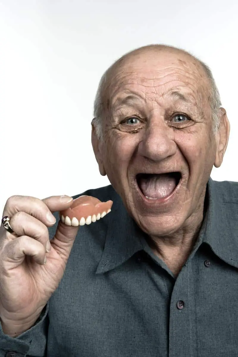 Elderly bald man without teeth wearing dark grey shirt with a huge open smile on his face holding dentures in his hand.