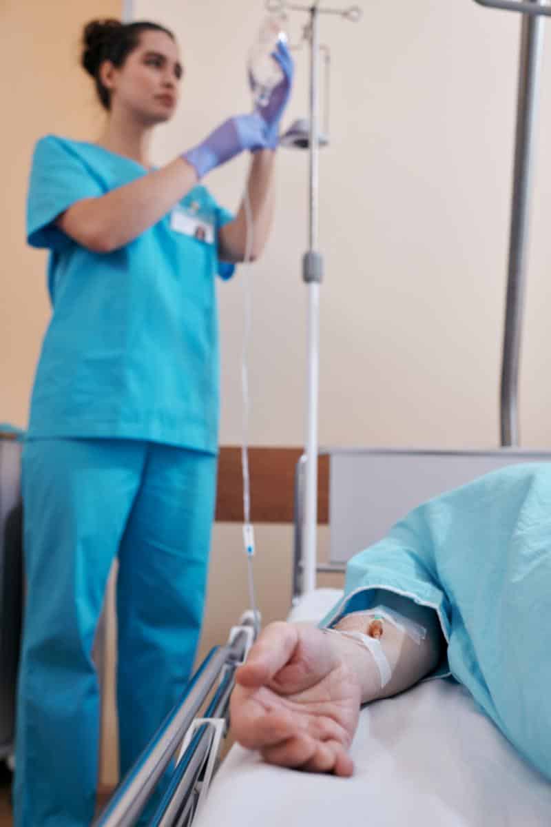 White female nursing with brown hair and teal scrubs wearing blue gloves hooking up IV fluids to patient laying in a hospital bed wearing a teal hospital gown. 