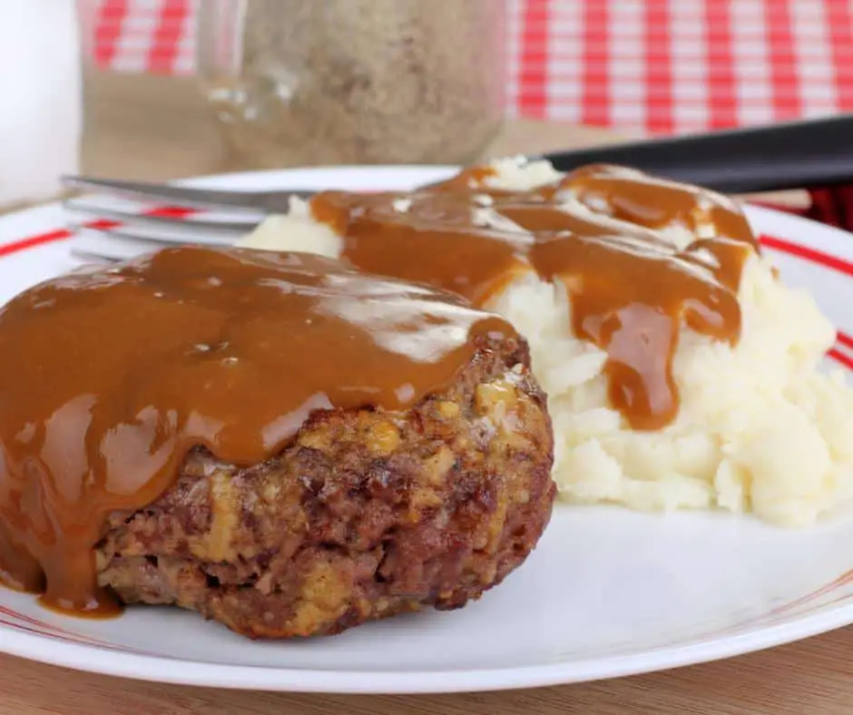 Meal to eat with no teeth: salisbury steak with gravy on top next to mashed potatoes with gravy on top of a white plate that has a red ring around it. 