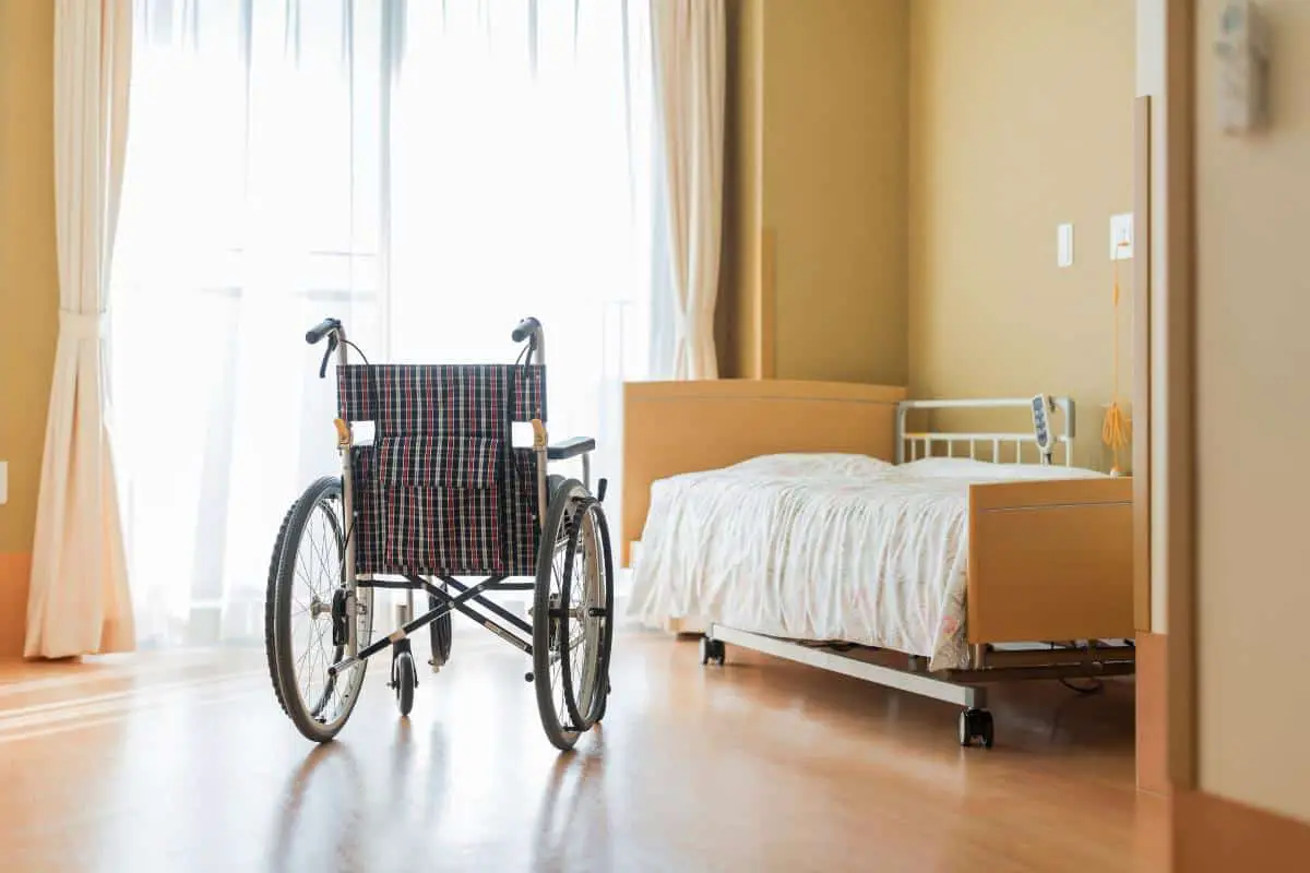 Empty wheelchair sitting in a facility room next to a hospital bed in front of a window with sunlight shining in.