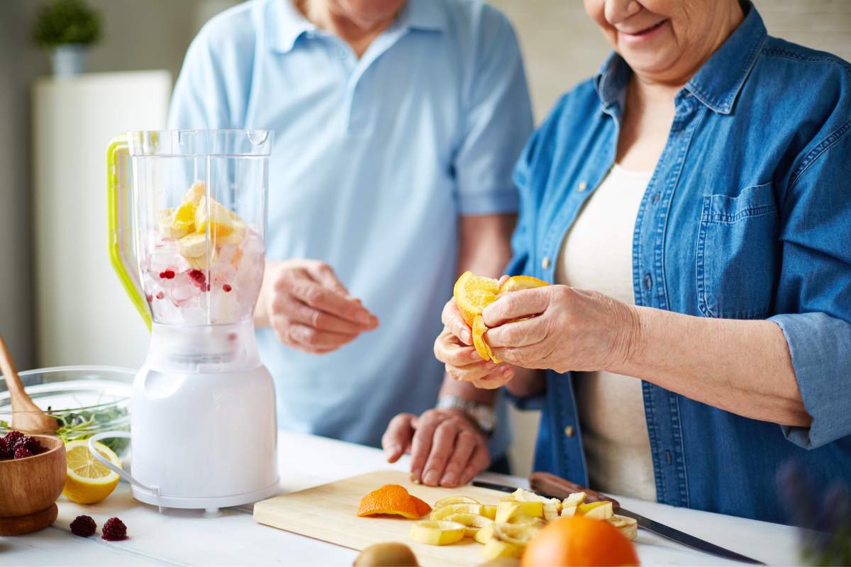 Older couple cutting up fruit and adding it to a blender to make nutritional drinks. 