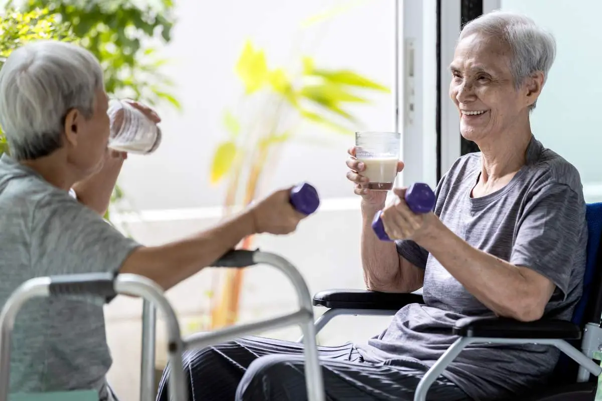 Two older adults sitting across from each other drinking nutritional drinks and lifting small weights.