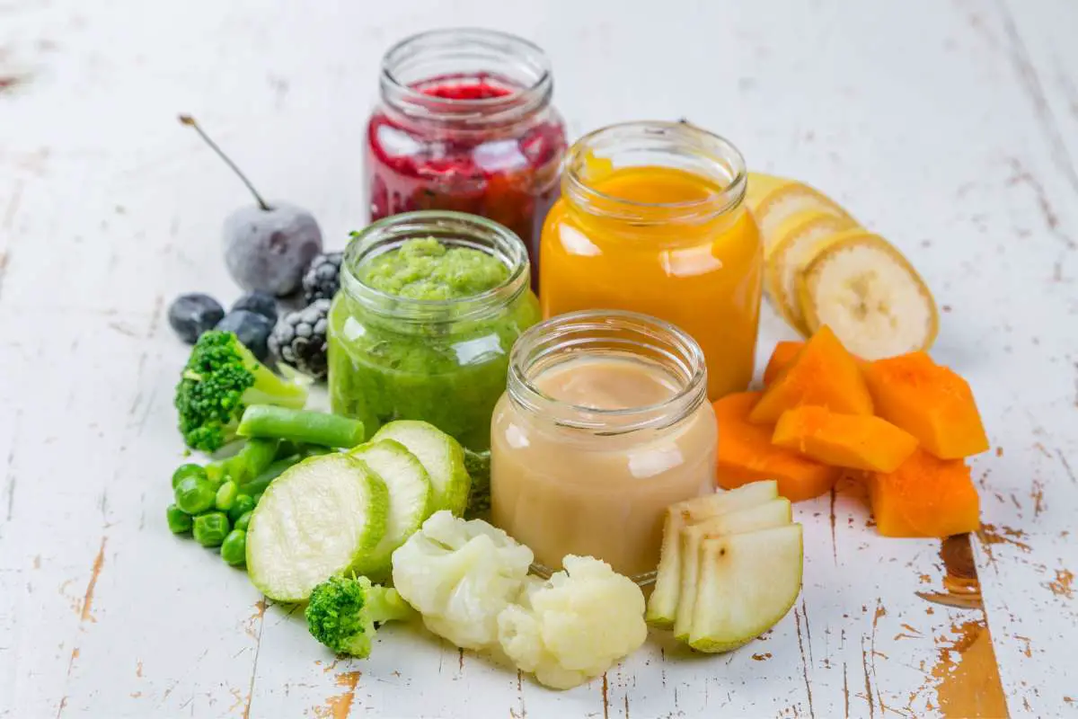 Whole berries, broccoli, zucchini, cauliflower, pears, carrots, bananas surrounding the same pureed items in clear jars sitting on grey background.